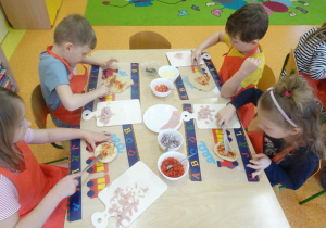 Czwórka dzieci smaruje sosem pomidorowym placek w kształcie koła przy użyciu noży.